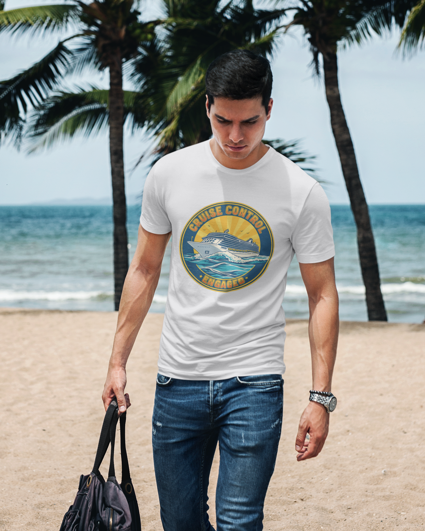 Man walking on a beach wearing a white t-shirt with a graphic design, holding a black bag.