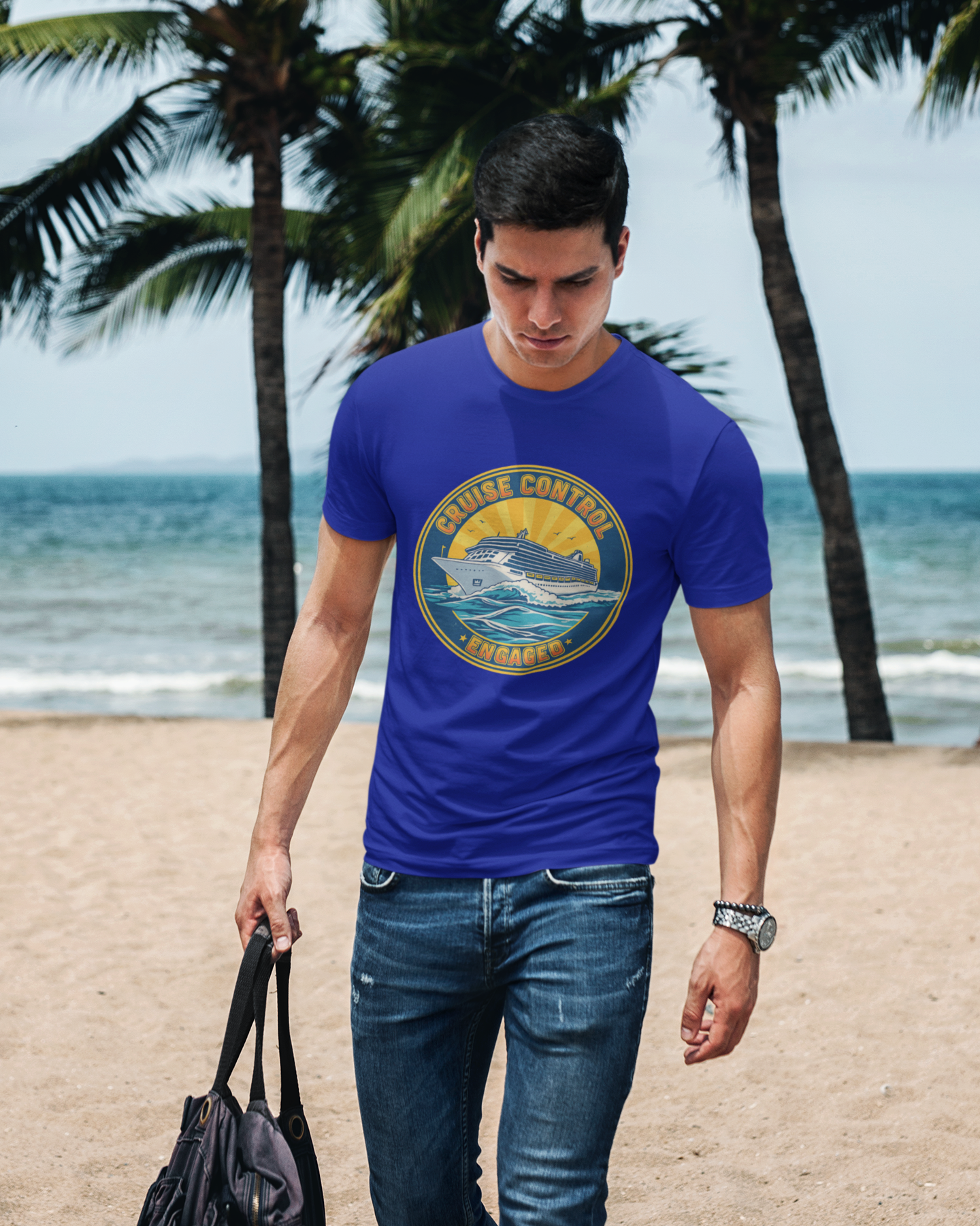 Man walking on a beach wearing a blue t-shirt with a logo, holding a black bag.