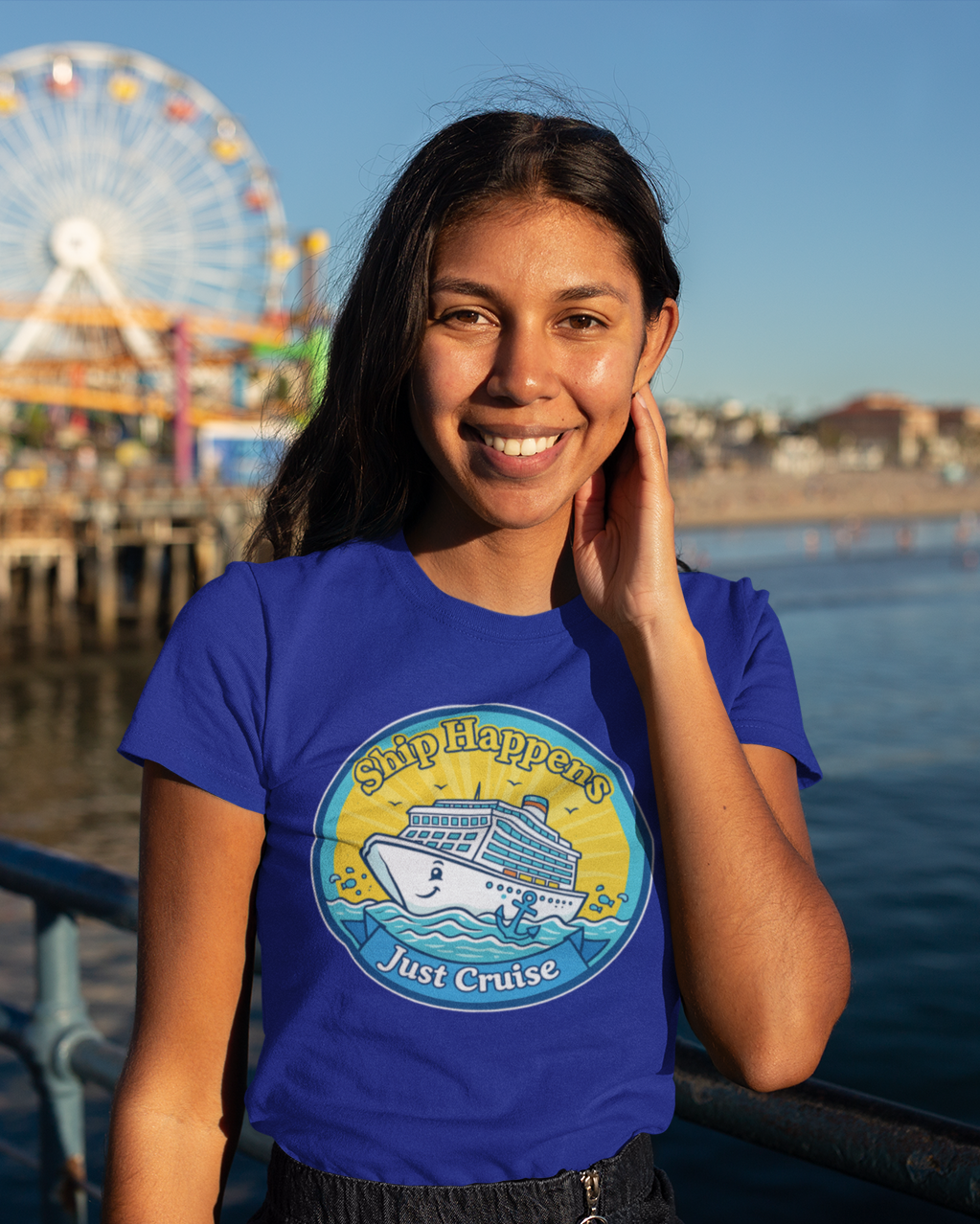 Woman wearing a blue t-shirt with a ship graphic and text, standing by water with a Ferris wheel in the background.