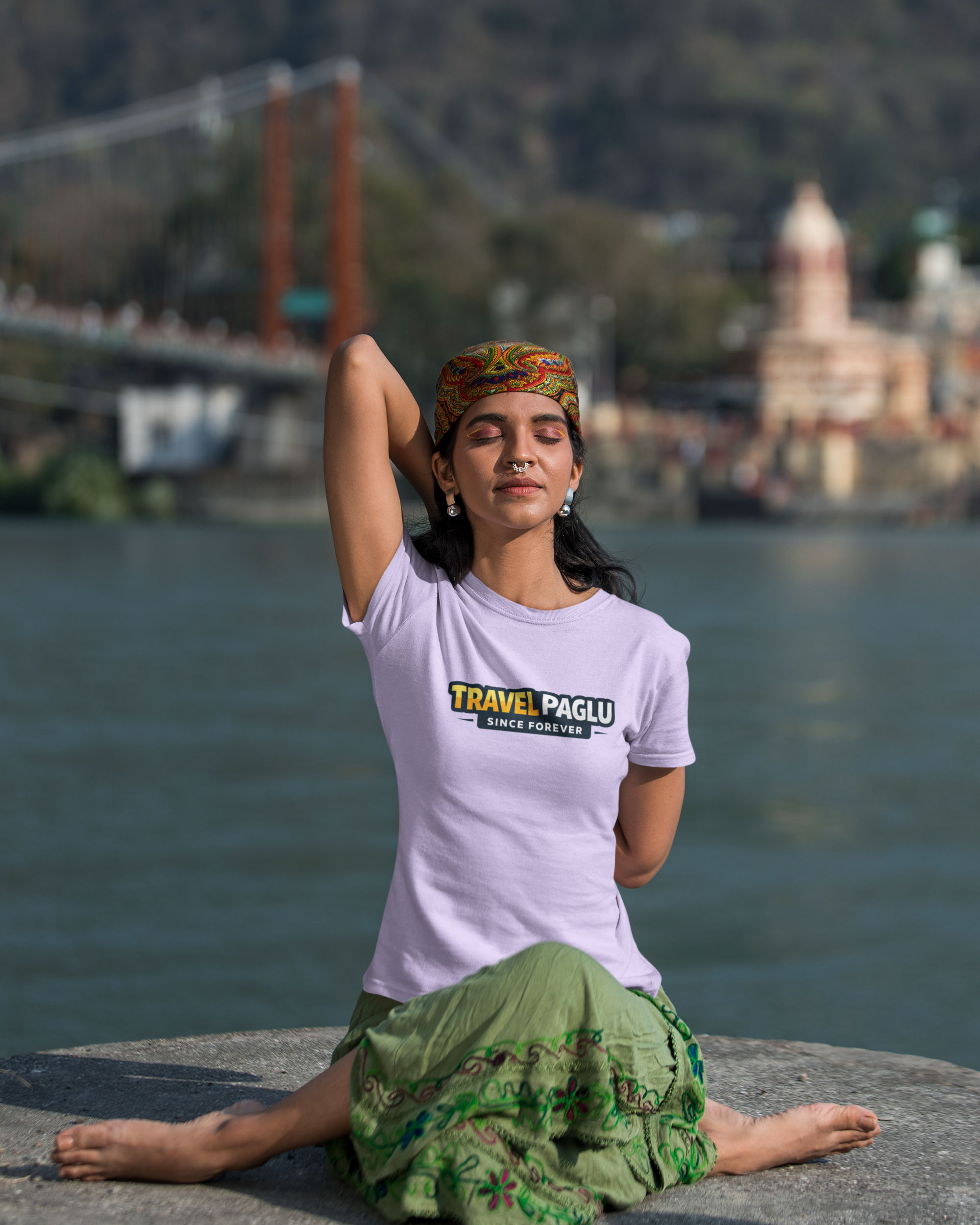 Woman in a white t-shirt with 'Travel Paglu' logo sitting by a body of water with mountains in the background.