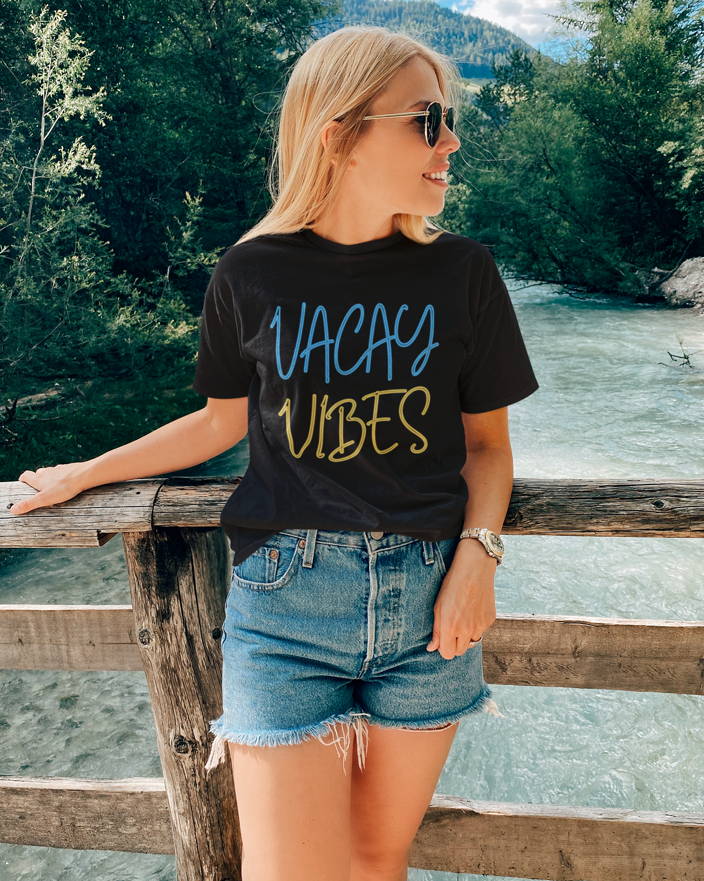 Woman wearing a black t-shirt with 'VACAY VIBES' text, standing by a river with greenery.