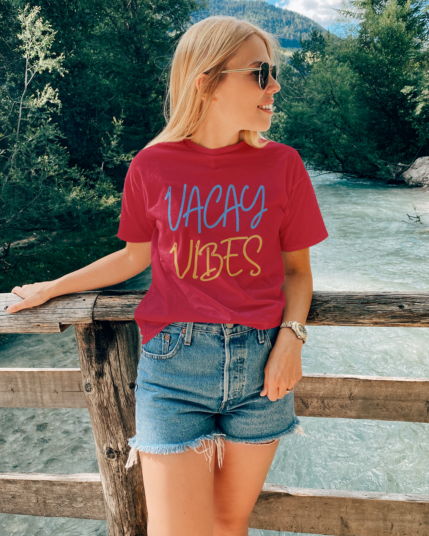 Woman wearing a red 'Vacay Vibes' t-shirt by a river with mountains in the background