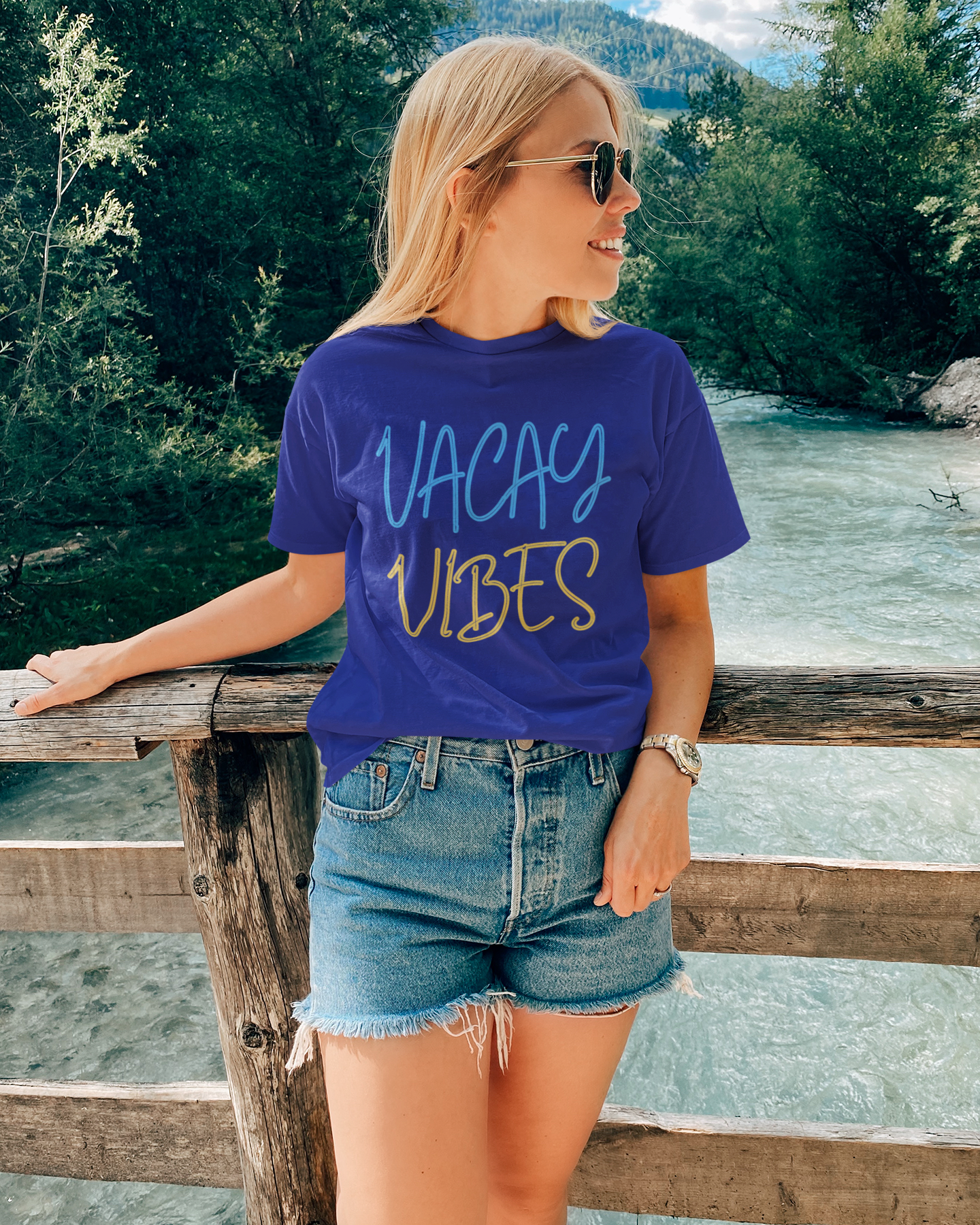 Woman wearing a blue 'VACAY VIBES' t-shirt by a river with mountains in the background