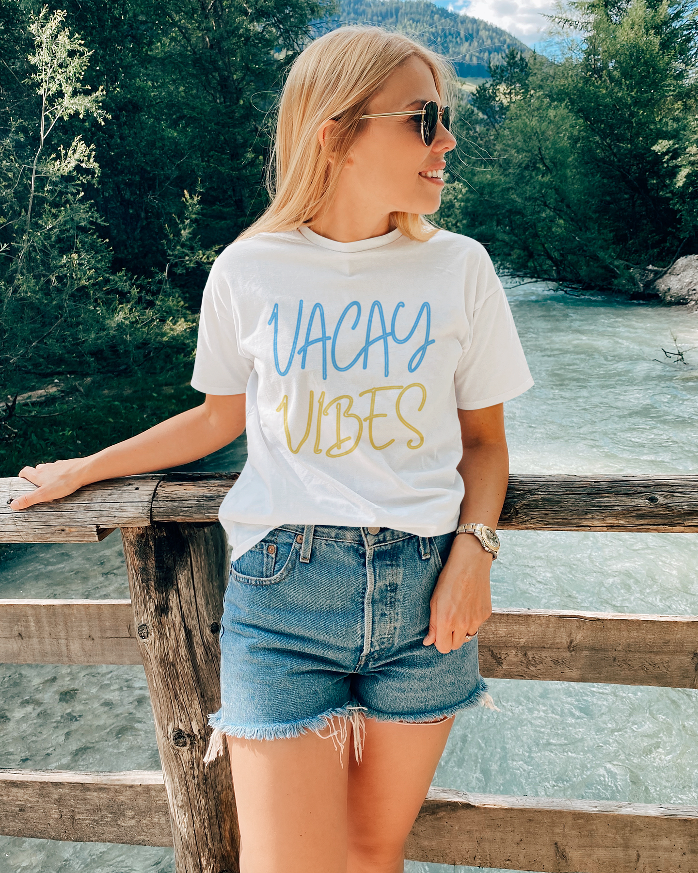Woman wearing a 'Vacay Vibes' t-shirt by a river with mountains in the background