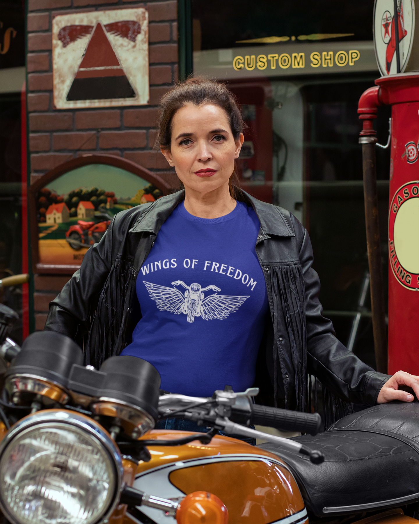 Woman wearing a blue 'Wings of Freedom' t-shirt standing next to a motorcycle in a custom shop setting.