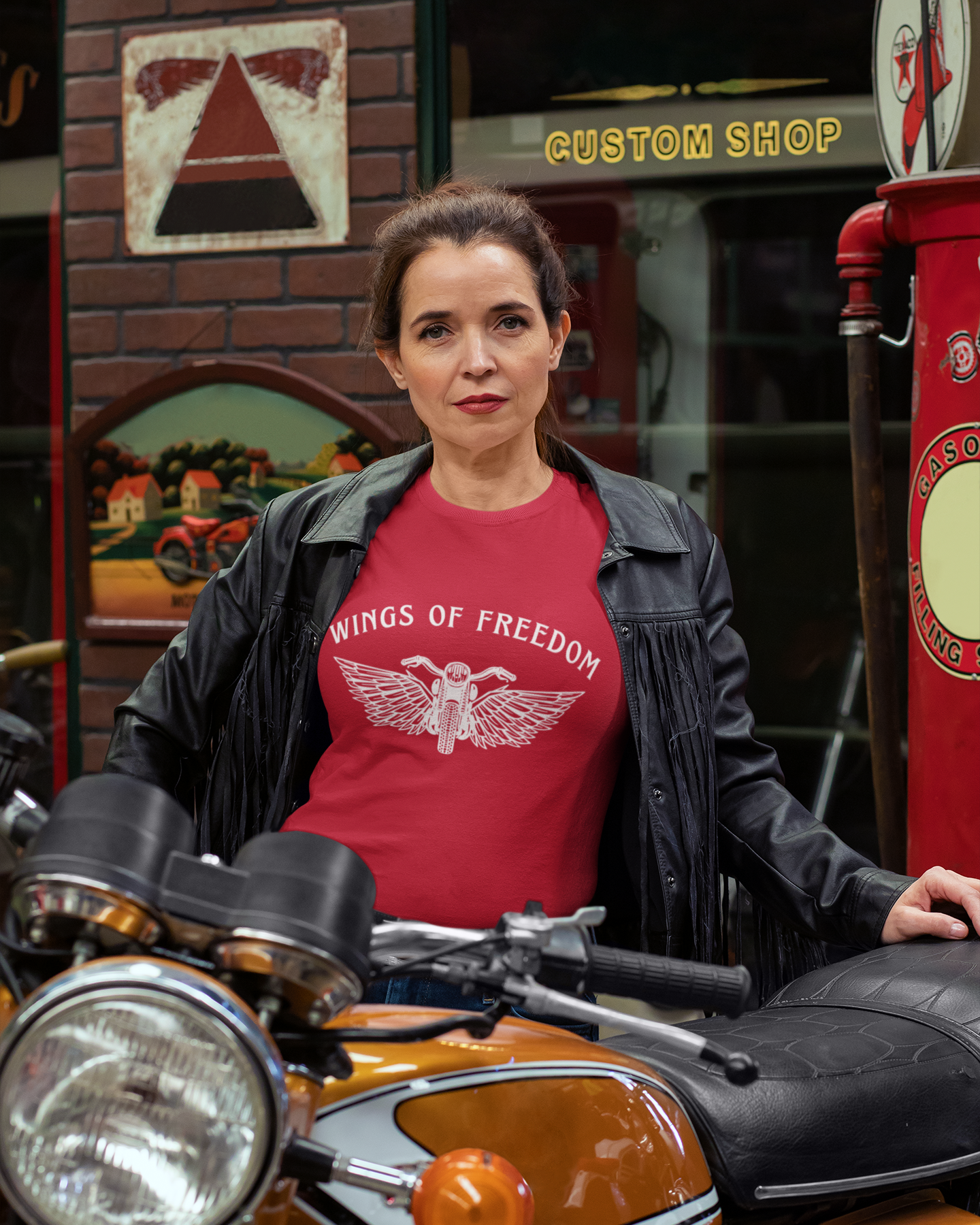 Woman wearing a red 'Wings of Freedom' t-shirt standing next to a motorcycle in a custom shop setting.