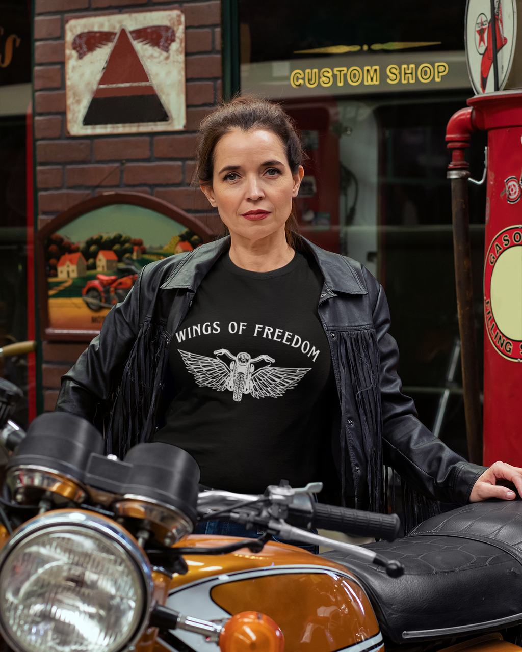 Woman wearing a 'Wings of Freedom' t-shirt sitting on a motorcycle in a custom shop setting.