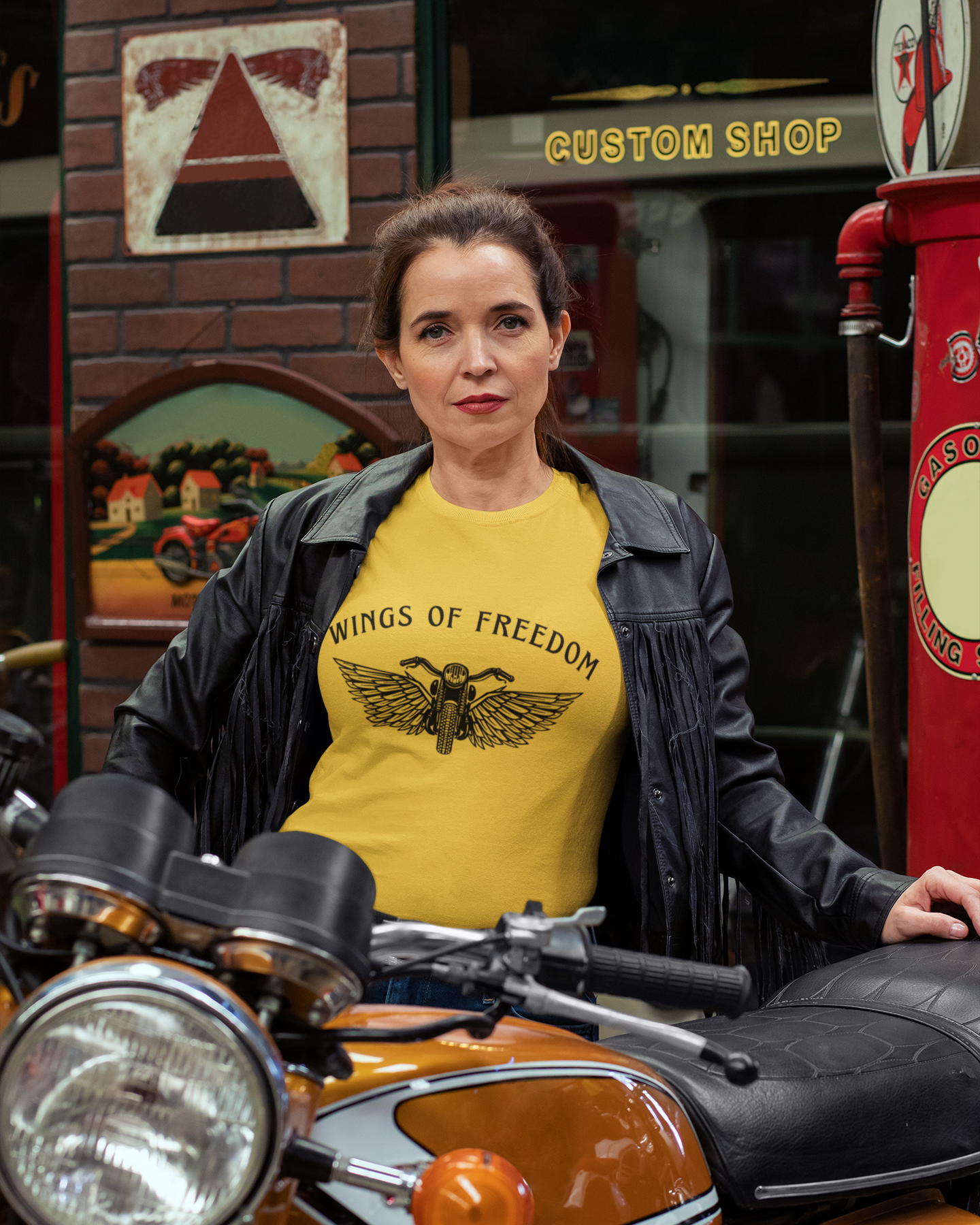 Woman wearing a yellow 'Wings of Freedom' t-shirt standing next to a motorcycle in a custom shop setting.