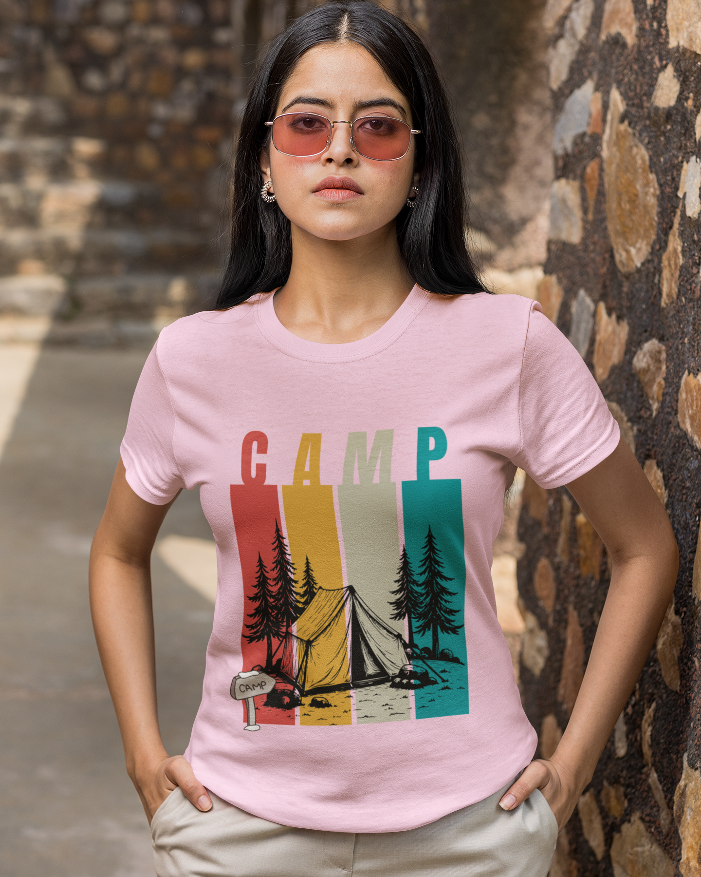 Person wearing a pink t-shirt with 'CAMP' and nature graphic, standing against a stone wall.