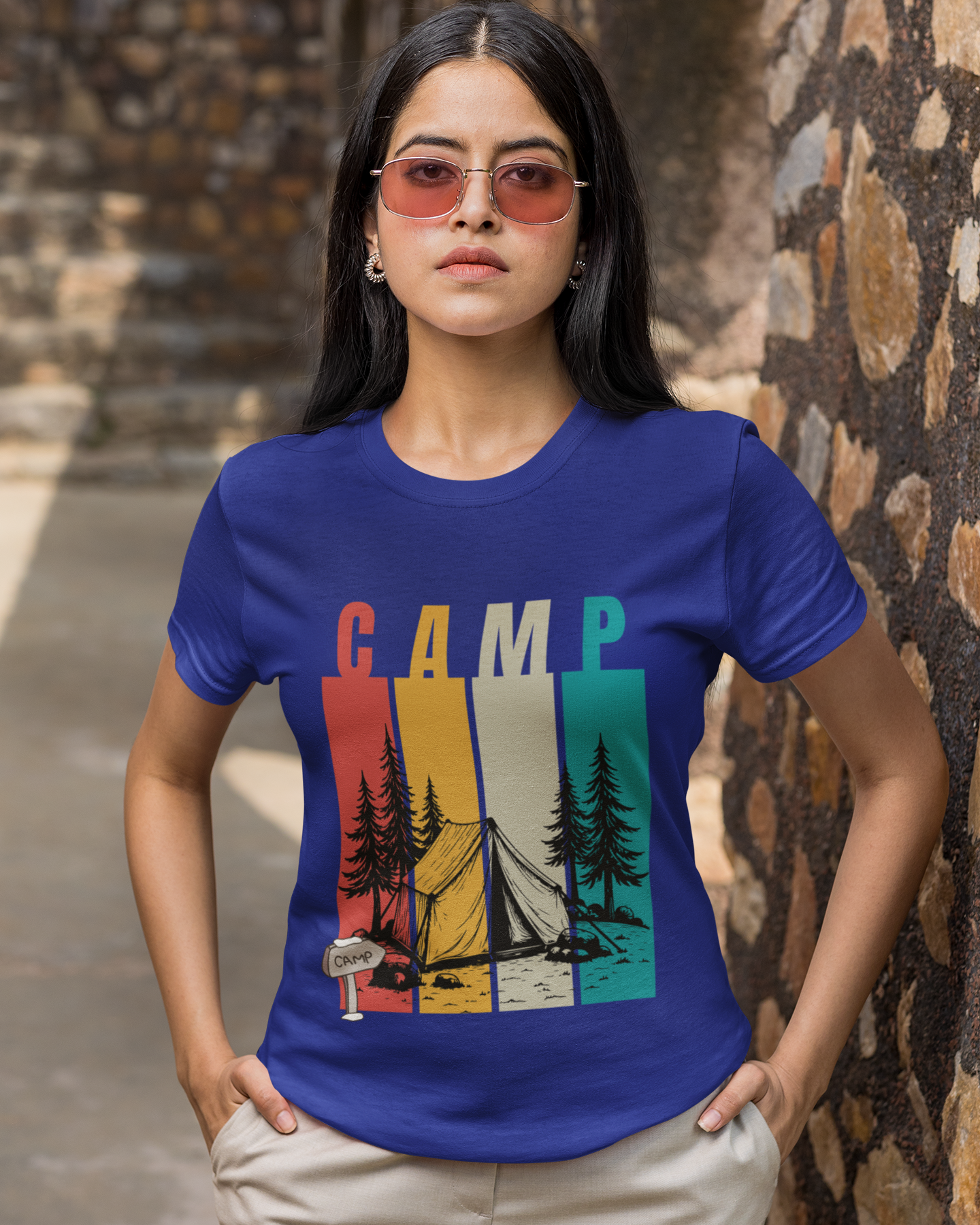 Woman wearing a blue t-shirt with 'CAMP' graphic design standing against a stone wall.