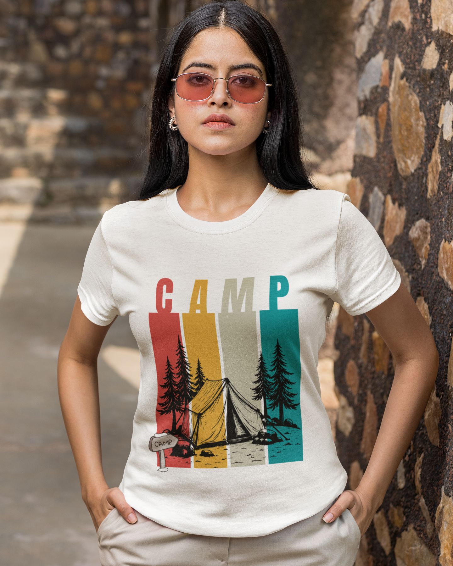 Woman wearing a white t-shirt with 'CAMP' graphic design standing against a stone wall.